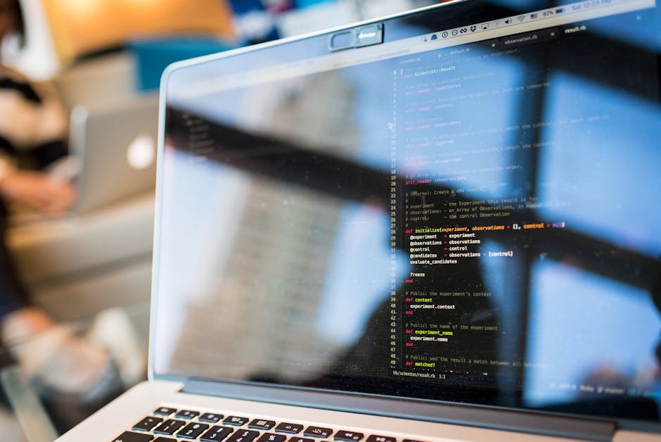 A close-up view of a laptop screen displaying lines of colorful programming code with syntax highlighting in shades of blue, pink, yellow, and white, situated on a desk in an indoor environment. In th