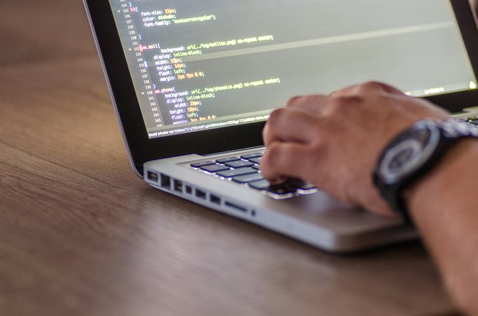 A close-up view of a laptop screen displaying lines of colorful programming code with syntax highlighting in shades of blue, pink, yellow, and white, situated on a desk in an indoor environment. In th