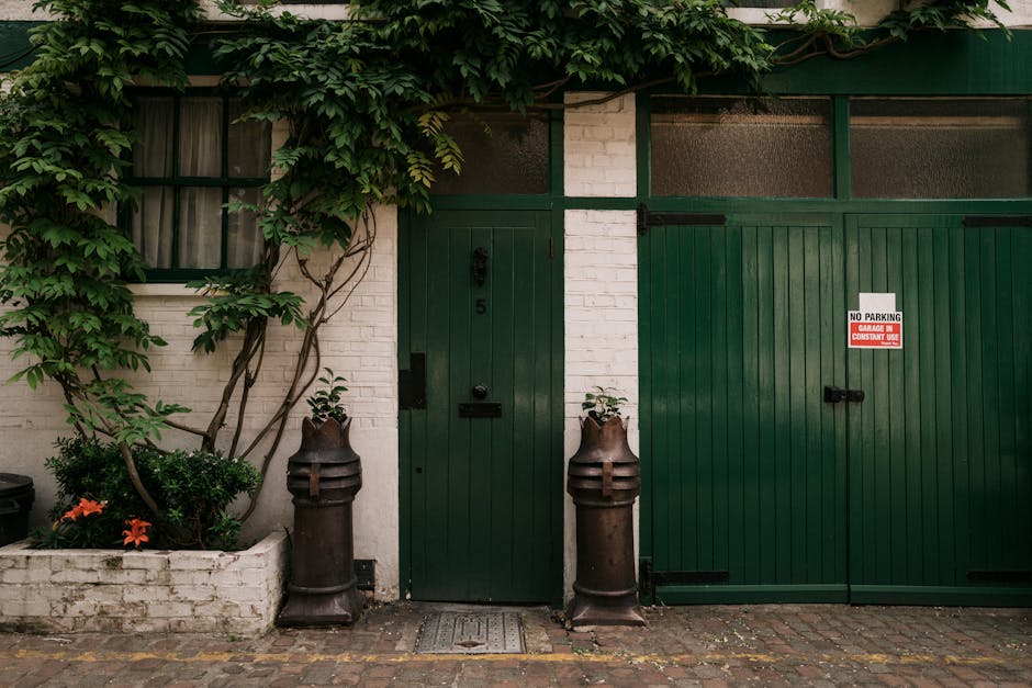 A view of a green wooden garage door and a matching pedestrian door set within a white brick wall, surrounded by lush green ivy and climbing plants. To the left of the pedestrian door, there is a smal